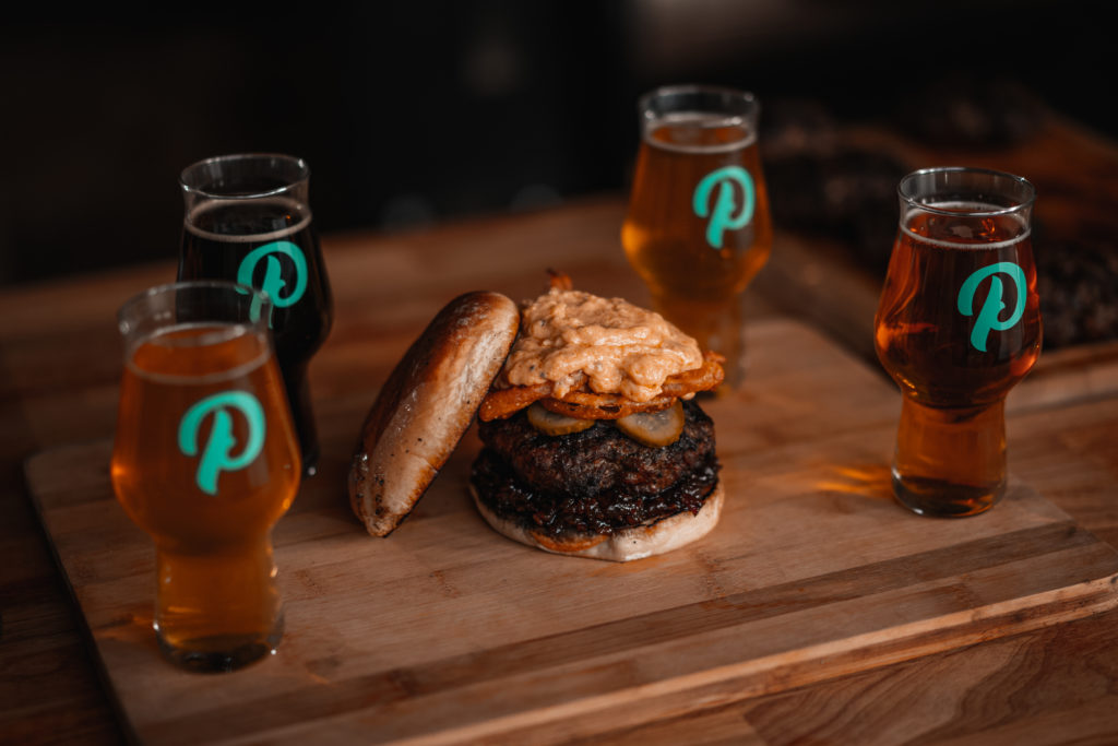 Product photography of a burger sitting in the middle of a beer flight with glasses displaying the Printshop Beer logo.