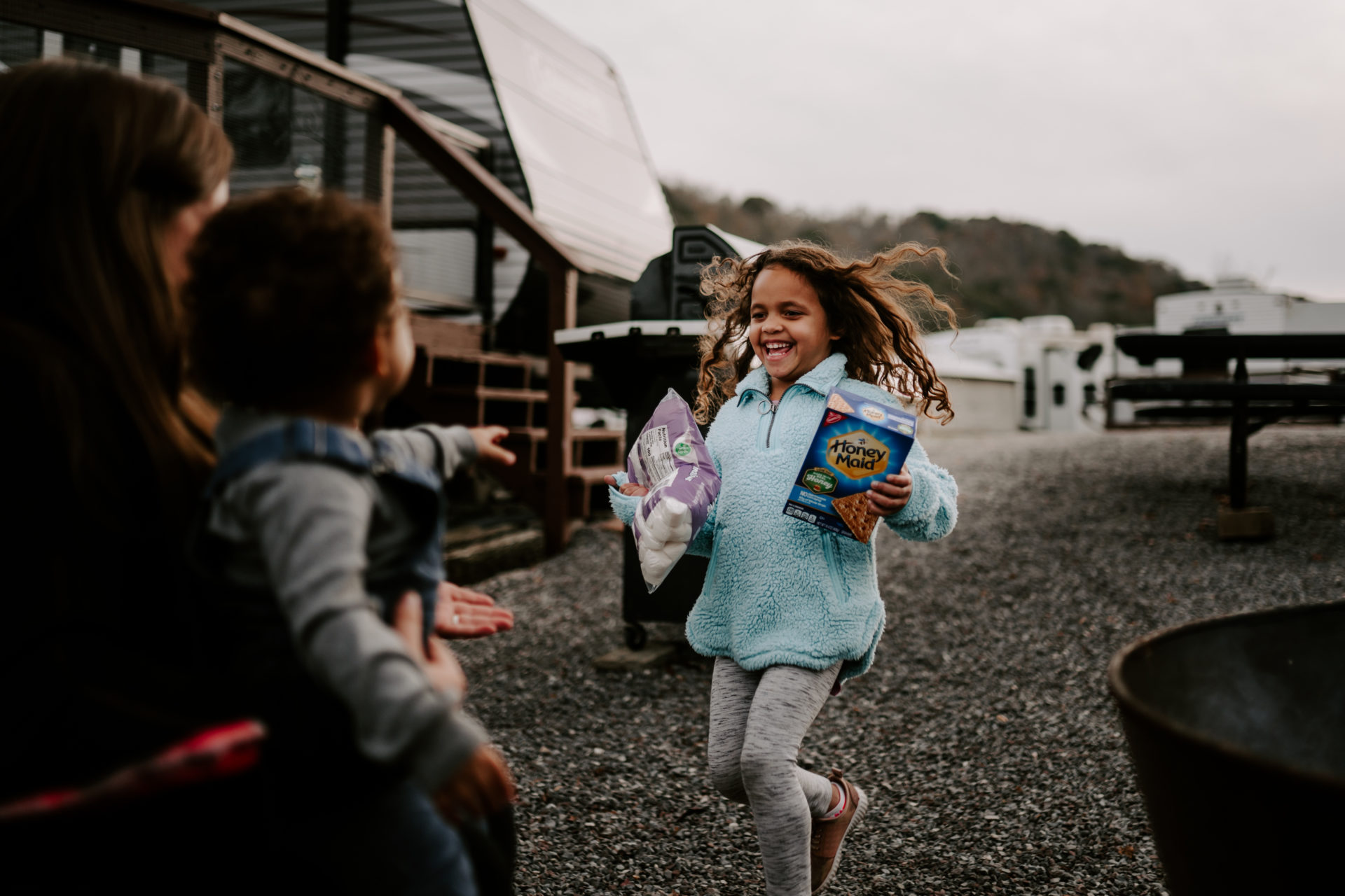 Lifestyle photography of a happy child running toward her mom while holding s'mores supplies in East TN.