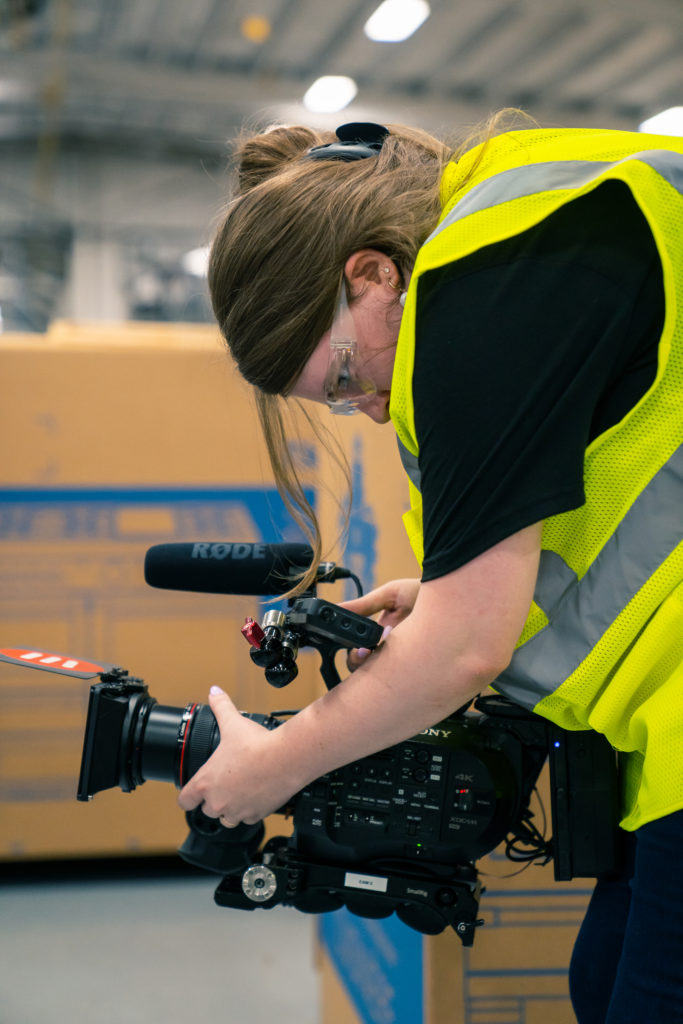 Megan Allen wearing a bright green work vest, pointing a camera and shooting in a warehouse