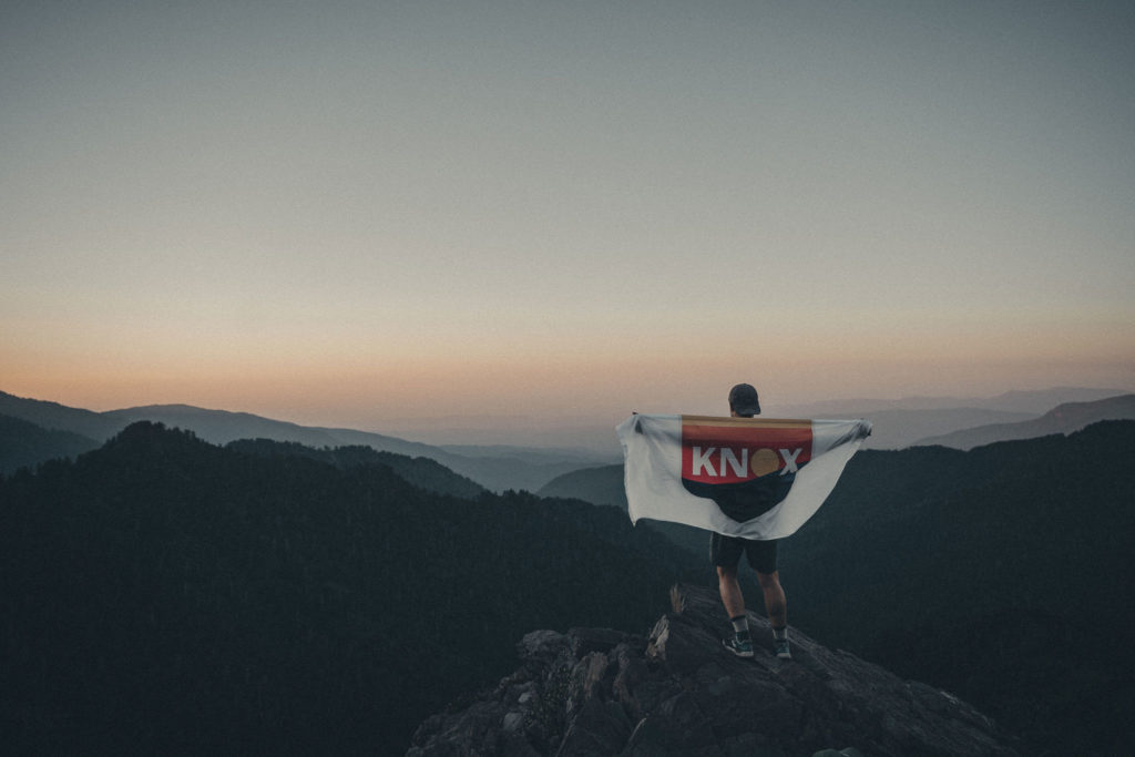 Lifestyle photography of a man standing on the top of a mountain overlooking an East Tennessee sunrise while holding a OneKnox flag over his shoulders.