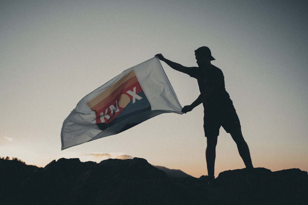 Lifestyle photography of a man standing on the top of a mountain overlooking East Tennessee while holding a OneKnox flag in the wind.