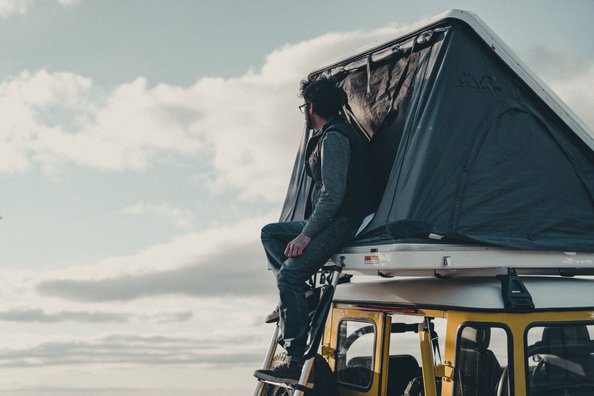 Commercial Photography of a man sitting along the outside of a rooftop tent on top of a car looking out into the distance.