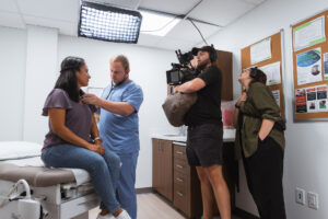 A doctor in blue scrubs uses a stethoscope to examine a female patient sitting on an exam table while a professional film crew records the scene in a medical office.