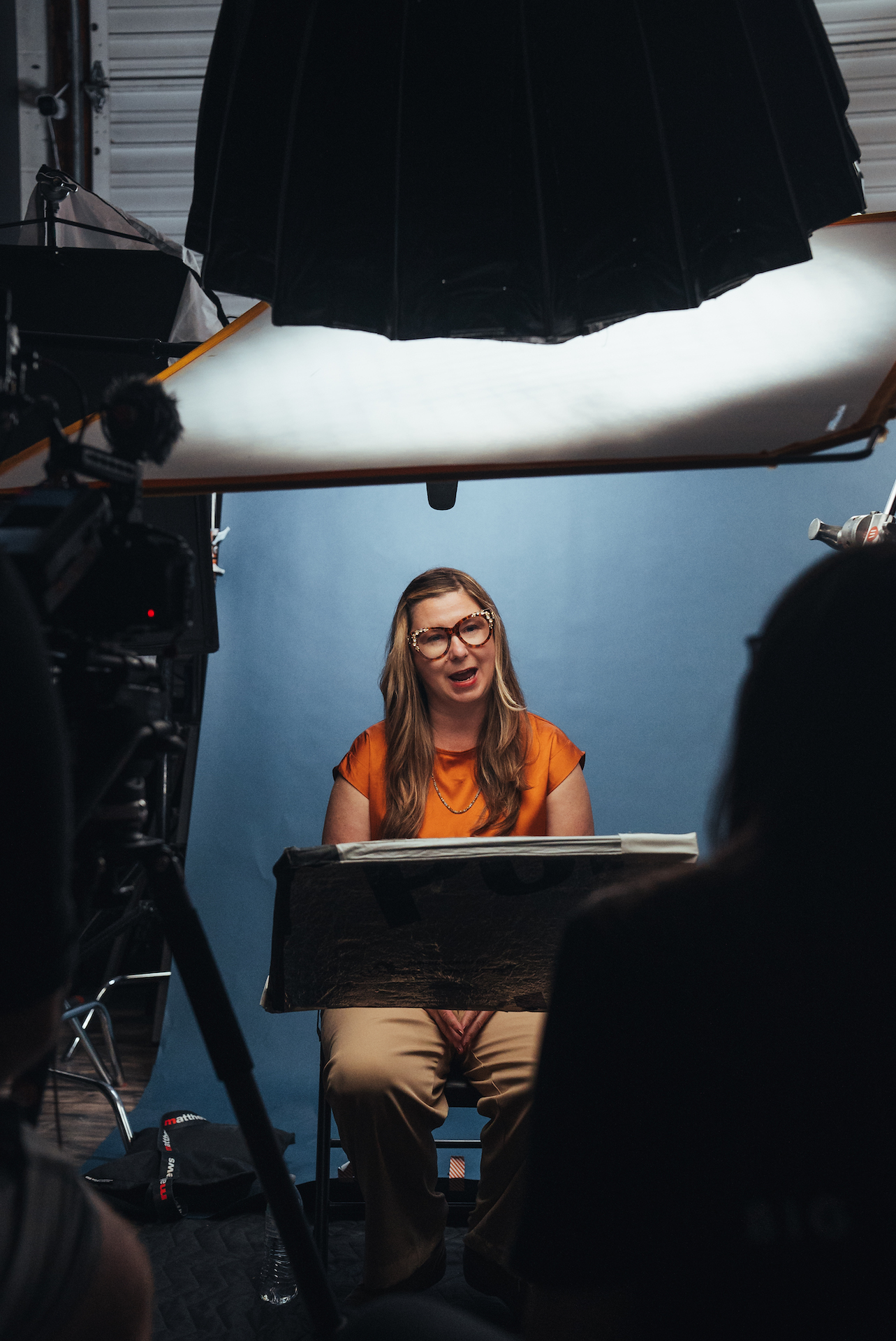 A woman in an orange top and glasses sits in front of a blue backdrop, speaking during a professionally lit video interview. Large softbox lighting and a camera crew surround the set, capturing the scene.