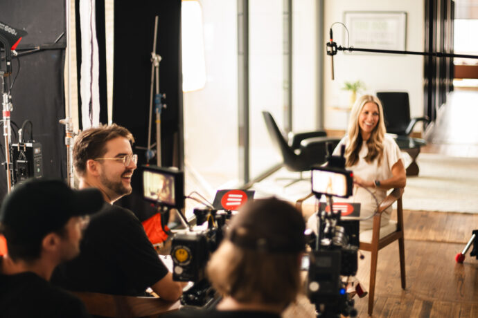 A behind-the-scenes view of a video production set, featuring a woman in a white outfit being interviewed while multiple cameras and a boom microphone capture the scene. The production crew, wearing black, operates the equipment in a bright, modern office space.