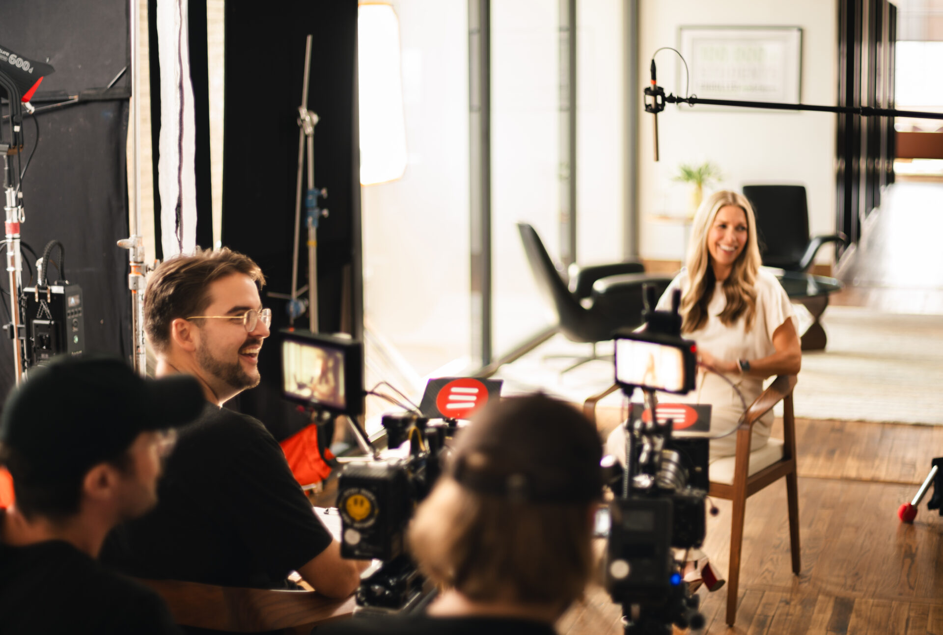 A behind-the-scenes view of a video production set, featuring a woman in a white outfit being interviewed while multiple cameras and a boom microphone capture the scene. The production crew, wearing black, operates the equipment in a bright, modern office space.