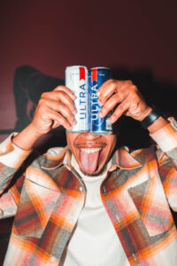 A smiling person in a plaid shirt playfully holds two Michelob Ultra cans over their eyes like goggles and sticks out their tongue, posing against a dark background.