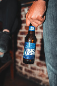 Close-up of a person holding a Bud Light beer bottle by the neck, standing against an exposed brick wall. Another person’s foot is visible in the background, resting on a bench.
