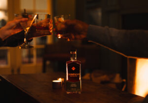Three people clink glasses of whiskey in a cozy, dimly lit setting with a fireplace. A bottle of Company Distilling bourbon sits on a wooden table in the foreground, next to a small candle.