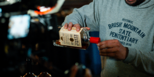 A person in a gray 'Brushy Mountain State Penitentiary' hoodie pours whiskey from a bottle labeled 'Double Barrel' at a dimly lit bar.