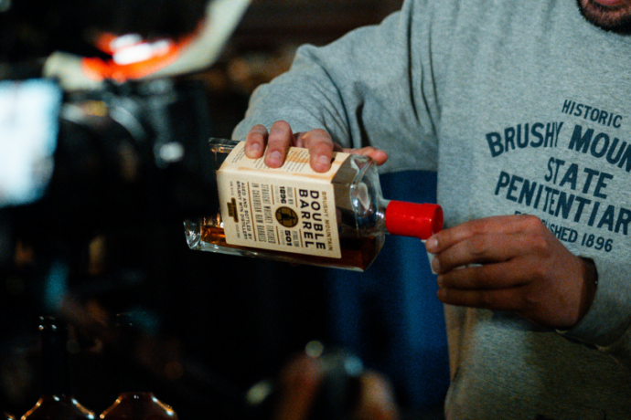 A person in a gray 'Brushy Mountain State Penitentiary' hoodie pours whiskey from a bottle labeled 'Double Barrel' at a dimly lit bar.