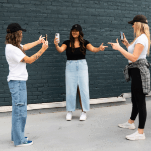social media intern Three women creating content together with smartphones in front of a dark brick wall; one stands in the center posing while the other two record her.