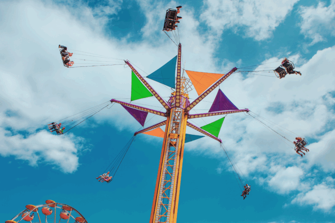 Colorful swing ride spinning high in the air at a fair, with riders flying against a bright blue sky with scattered clouds.