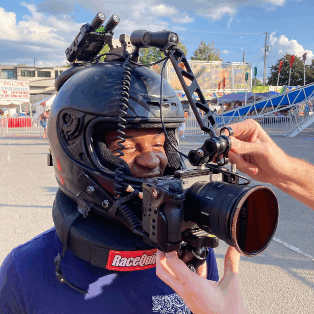 Man wearing a black helmet rigged with a mounted camera setup, preparing to film at a fairground with rides and food stalls in the background.