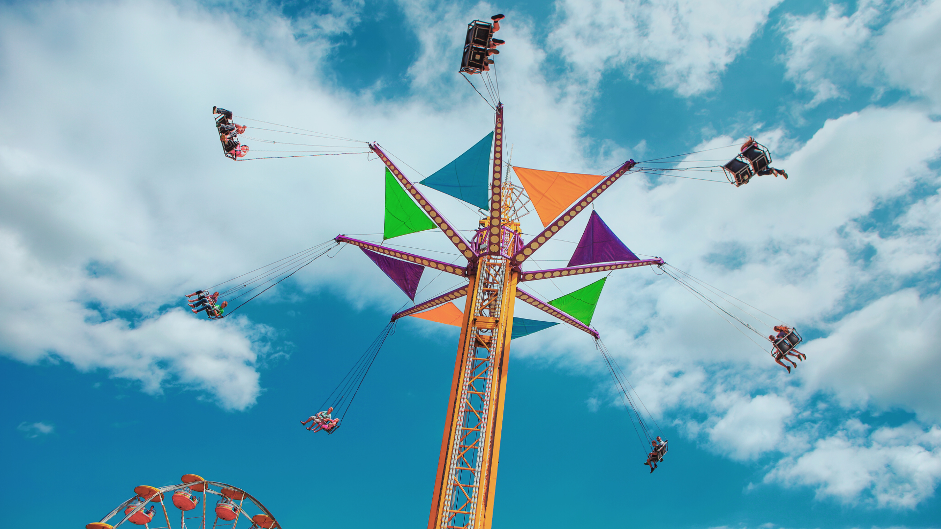 Colorful swing ride spinning high in the air at a fair, with riders flying against a bright blue sky with scattered clouds.