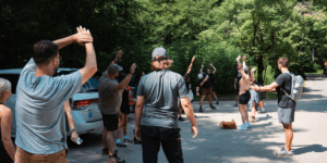 A group of people stretches together in a sunny parking area surrounded by trees, preparing for an outdoor activity. Some participants have backpacks and hiking gear, while a few cars are parked nearby. The atmosphere appears casual and energetic.