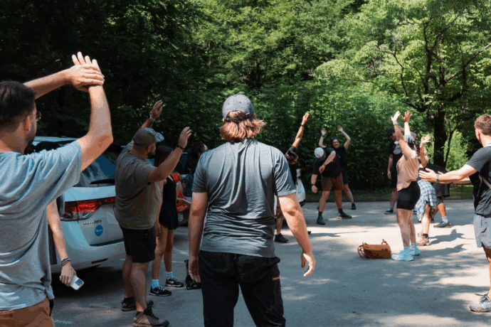 A group of people stretches together in a sunny parking area surrounded by trees, preparing for an outdoor activity. Some participants have backpacks and hiking gear, while a few cars are parked nearby. The atmosphere appears casual and energetic.