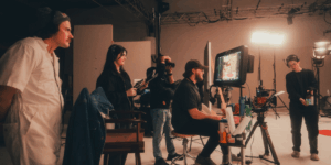 Behind-the-scenes view of a production team on set, gathered around a monitor displaying the live camera feed. Some team members are wearing headsets and holding equipment, while professional lighting and filming gear fill the studio space. The atmosphere is focused and collaborative.