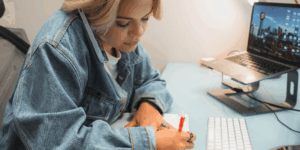 A woman wearing a denim jacket working at a social media agency multitasks at her desk, holding a phone between her ear and shoulder while writing notes with a red pen. A laptop and keyboard are on the desk in front of her.