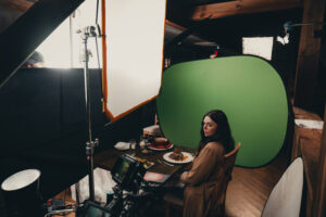 A behind-the-scenes photo of a video production setup featuring a woman sitting at a restaurant table with plated food in front of her. She is positioned in front of a green screen, surrounded by professional lighting equipment, camera gear, and crew members. The scene is lit with softboxes and reflectors, creating a controlled environment for compositing or visual effects.