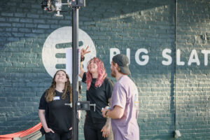 Three members of KnoxMedia smile and look up while adjusting a camera rig outside the Big Slate Media building, with a branded mural visible in the background.