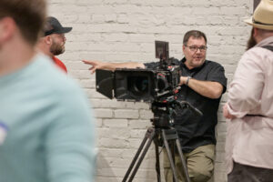A man at the KnoxMedia meet-up gestures while discussing a RED cinema camera setup on a tripod with two other attendees indoors, standing in front of a white brick wall.
