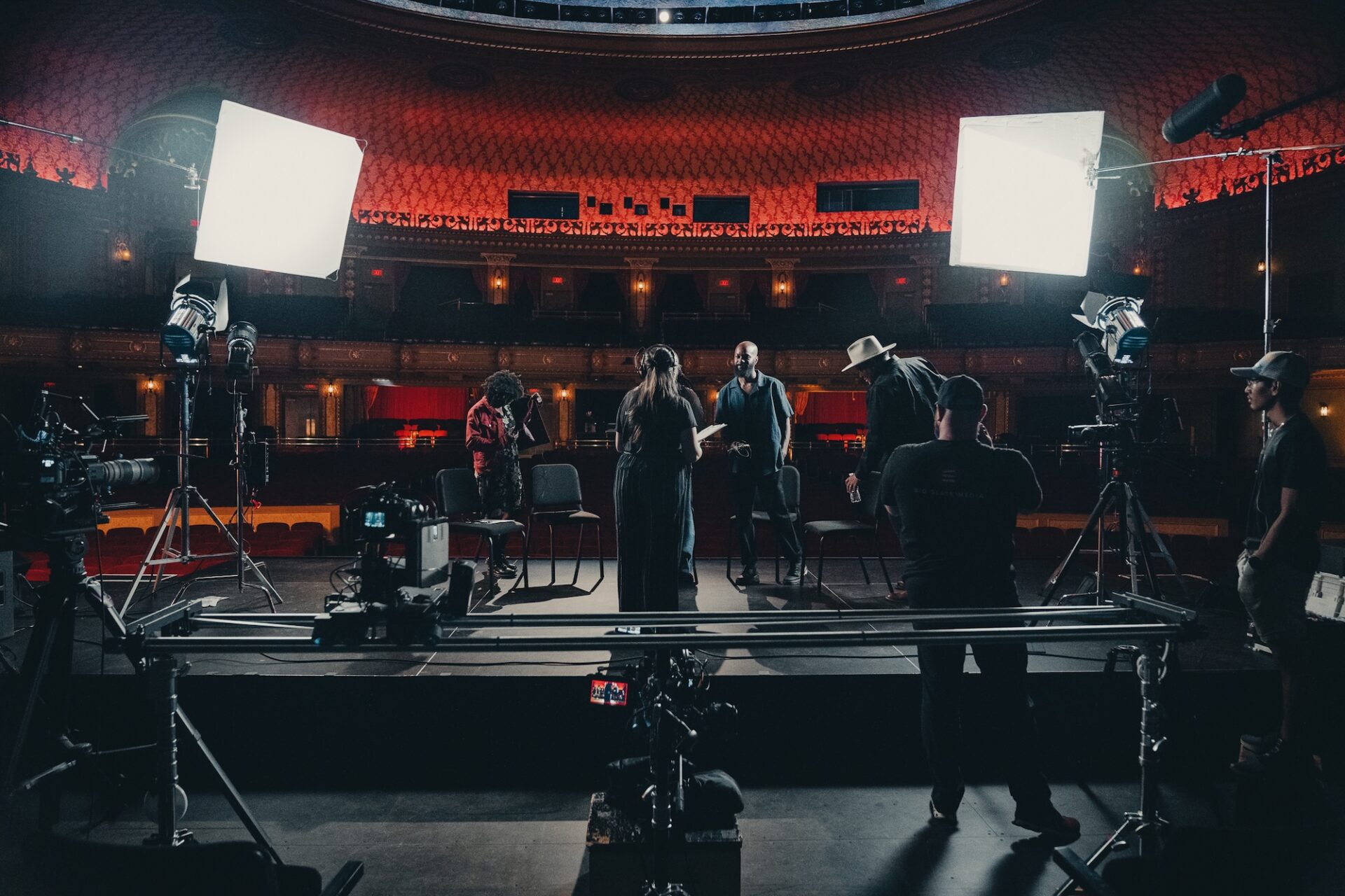 A behind-the-scenes view of a video shoot on the Tennessee Theatre stage. Crew members adjust lighting and cameras around a circle of chairs, preparing for a group interview. The grand interior of the historic theatre glows warmly in the background, adding depth and elegance to the setup.