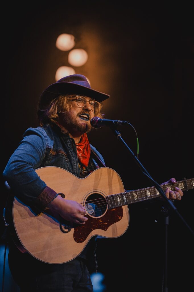 Adeem the Artist performs on stage at the Tennessee Theatre, playing an acoustic guitar and singing into a microphone. They wear a wide-brimmed hat, denim jacket, red bandana, and blue lipstick, with warm stage lights glowing in the background.