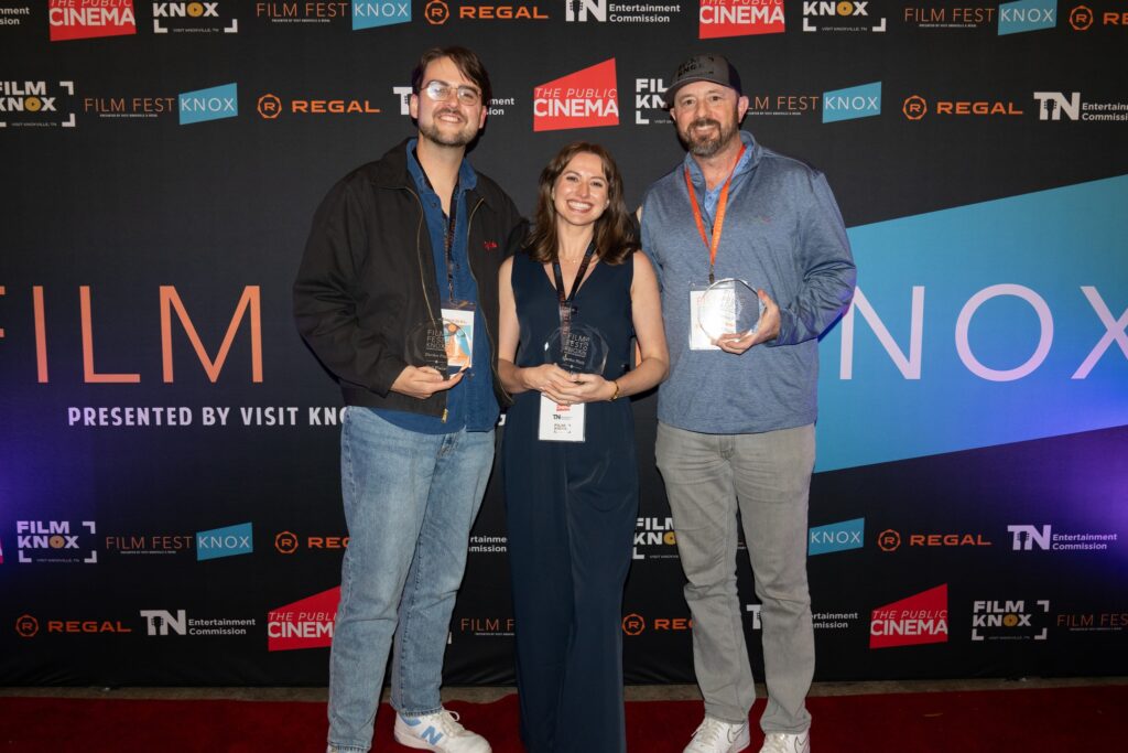 Three people stand together smiling on a red carpet in front of a Film Fest Knox backdrop featuring sponsor logos. Each person holds an award, and all are wearing festival lanyards around their necks.