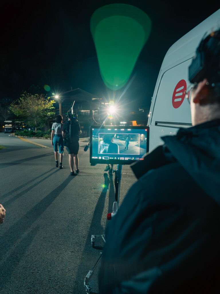A nighttime outdoor film set where a crew member watches a monitor showing a shot in progress. Ahead, two crew members walk with a performer toward a bright light source, casting long shadows on the pavement. A production van is parked to the right, and lens flares and set lights illuminate the scene.