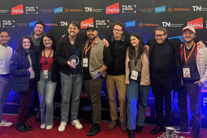 A group of ten people stand together smiling on a red carpet in front of a Film Fest Knox step-and-repeat backdrop featuring logos from The Public Cinema, Regal, and the Tennessee Entertainment Commission. One person in the center holds a clear award, and several wear festival lanyards around their necks.