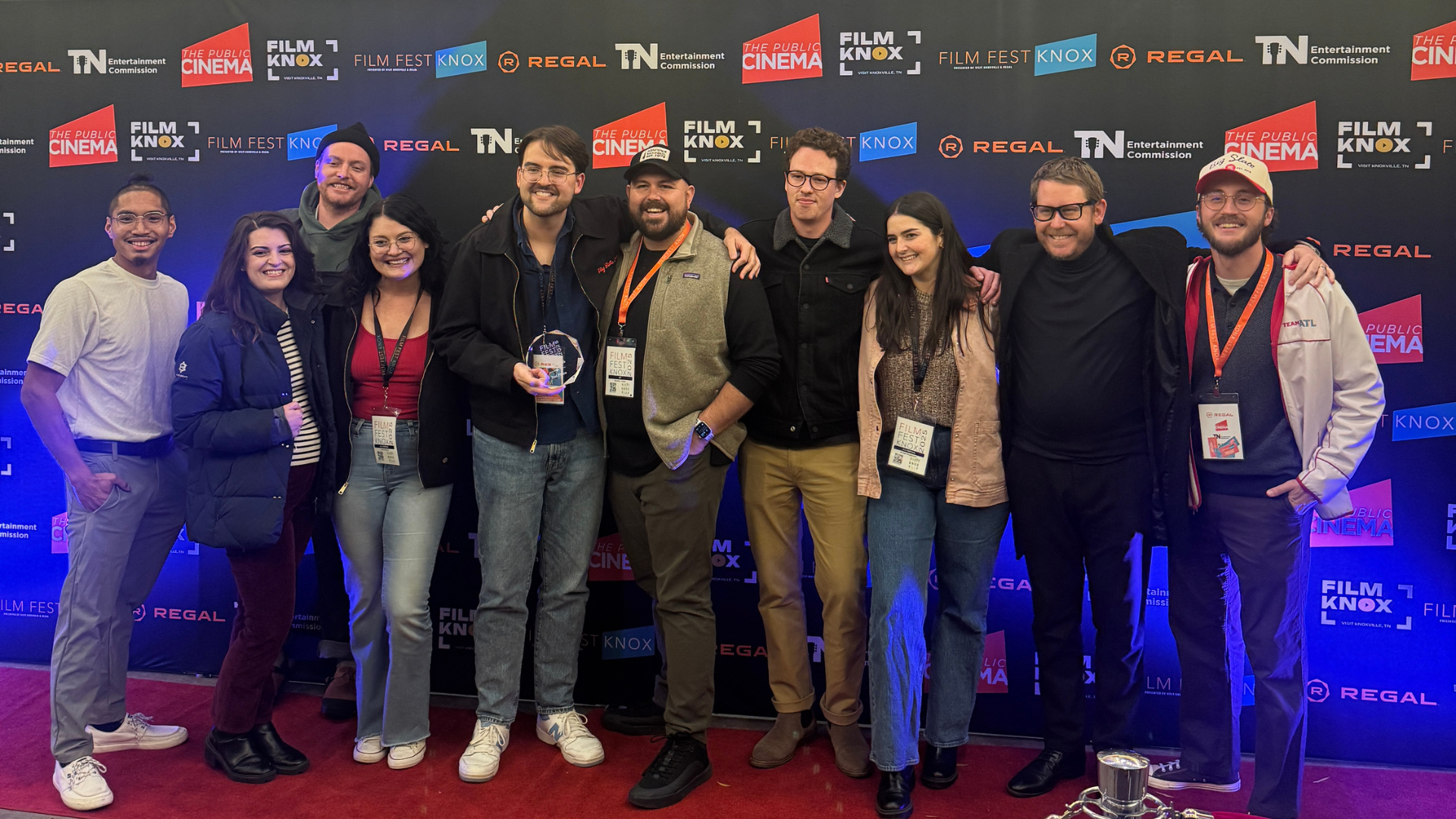 A group of ten people stand together smiling on a red carpet in front of a Film Fest Knox step-and-repeat backdrop featuring logos from The Public Cinema, Regal, and the Tennessee Entertainment Commission. One person in the center holds a clear award, and several wear festival lanyards around their necks.