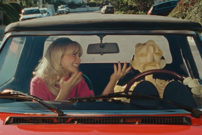 Sabrina Carptenter smiles and gestures while riding in a red convertible with a humanoid figure made of stacked Pringles driving; the scene is viewed through the windshield on a sunny residential street.