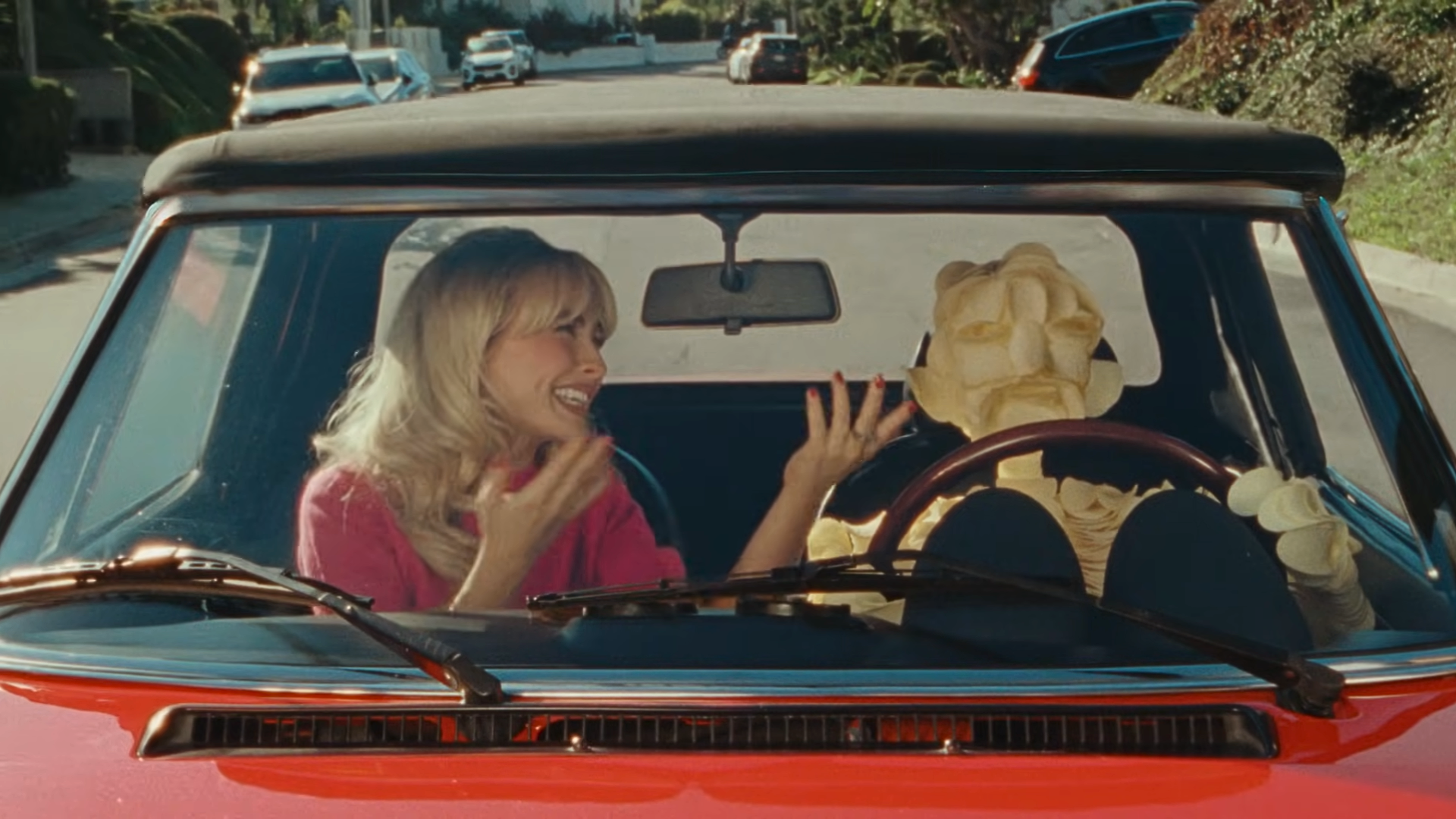 Sabrina Carptenter smiles and gestures while riding in a red convertible with a humanoid figure made of stacked Pringles driving; the scene is viewed through the windshield on a sunny residential street.