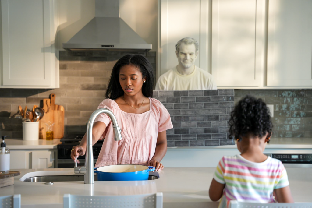A woman fills a pot with water in a bright modern kitchen while a child stands nearby, as a camouflaged man blends into the light-colored cabinetry behind them.