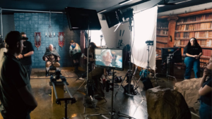 Wide behind-the-scenes view of a commercial production set with crew members, cameras, lighting equipment, and monitors surrounding an older actor seated in a medieval-inspired scene.