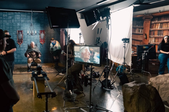 Wide behind-the-scenes view of a commercial production set with crew members, cameras, lighting equipment, and monitors surrounding an older actor seated in a medieval-inspired scene.