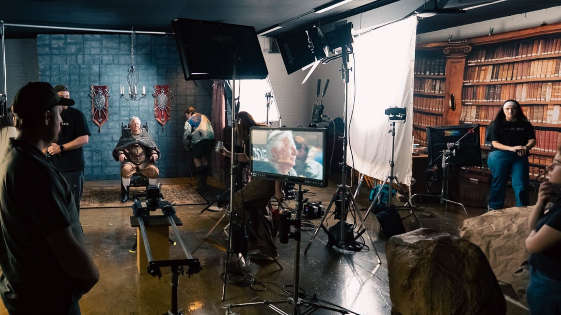 Wide behind-the-scenes view of a commercial production set with crew members, cameras, lighting equipment, and monitors surrounding an older actor seated in a medieval-inspired scene.