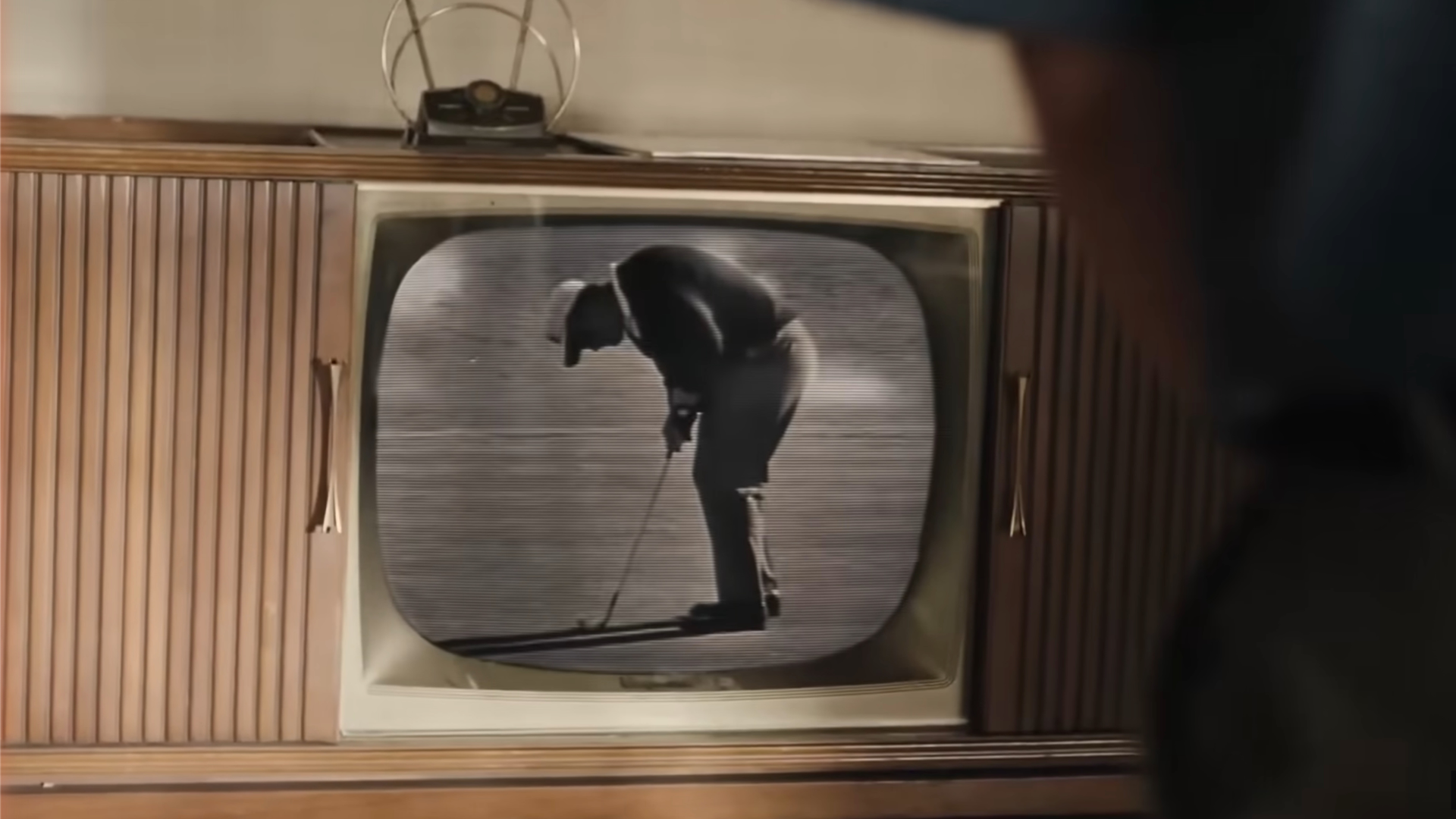 A vintage wooden television displays black-and-white footage of a golfer putting on a green, with part of a person visible in the foreground watching the screen.